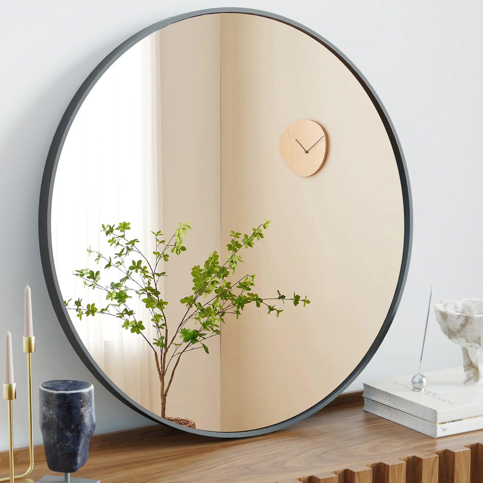 A black circular wall mirror hanging on a wall, reflecting a plant and a wooden clock.