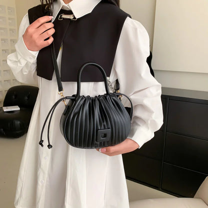 A person wearing a white dress and black vest holding a black PU leather bucket bag with a drawstring closure.