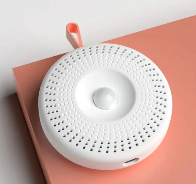 A white cat basin deodorizer with a round shape and a central control button, featuring small holes for ventilation on the surface, placed on a peach-colored surface.