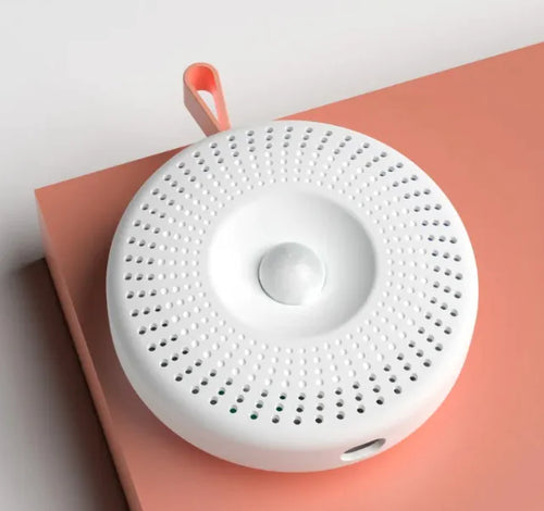 A white cat basin deodorizer with a round shape and a central control button, featuring small holes for ventilation on the surface, placed on a peach-colored surface.