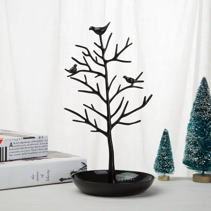 A gold-colored metal jewelry display stand with a bird motif on top, featuring a dish at the base for holding jewelry items, displayed next to a white cup and saucer on a wooden surface.