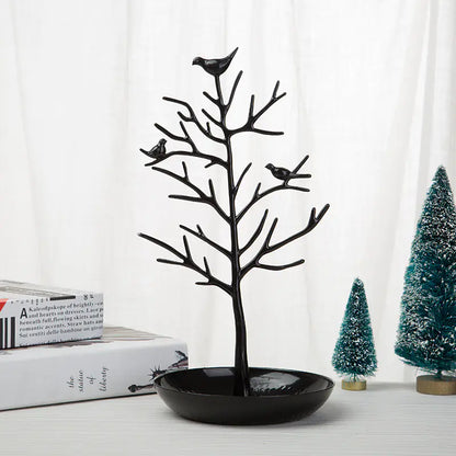 A gold-colored metal jewelry display stand with a bird motif on top, featuring a dish at the base for holding jewelry items, displayed next to a white cup and saucer on a wooden surface.