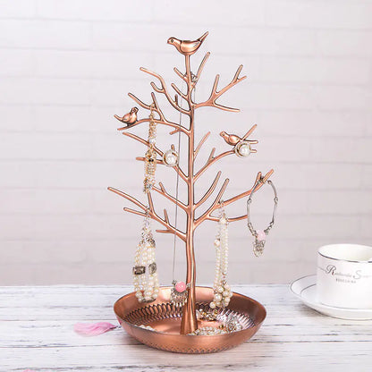 A gold-colored metal jewelry display stand with a bird motif on top, featuring a dish at the base for holding jewelry items, displayed next to a white cup and saucer on a wooden surface.
