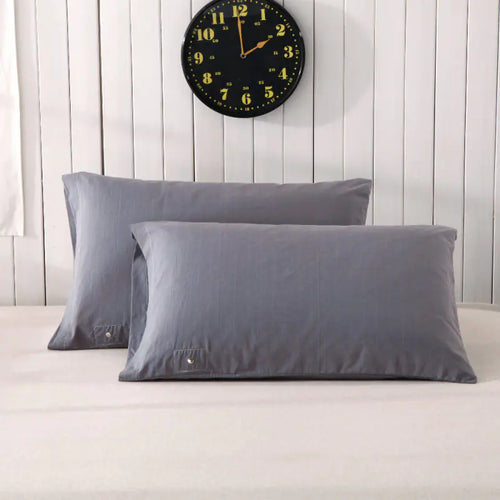 Two gray pillowcases on a bed with a white sheet, against a wooden wall with a clock above. The pillows are positioned at an angle, showcasing the brand tag on one of them.