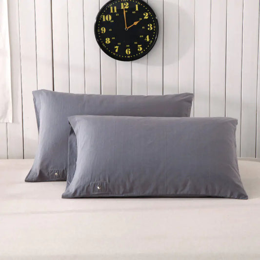 Two gray pillowcases on a bed with a white sheet, against a wooden wall with a clock above. The pillows are positioned at an angle, showcasing the brand tag on one of them.