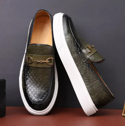 A black casual shoe with a crocodile pattern, featuring a metal embellishment on the toe and a flat sole, placed on a wooden surface next to a brass object.