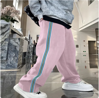 A pair of men's casual trousers in pink, with a striped pattern featuring green and white lines on the side, worn with a grey sweater and white sneakers.