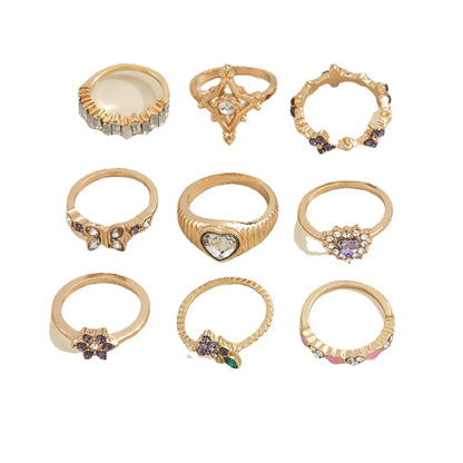 A collection of gold-colored geometric rings with purple and clear accents on a hand with white nails.