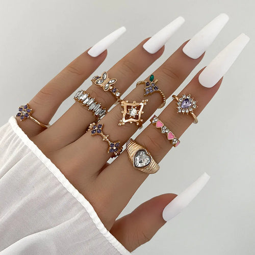 A collection of gold-colored geometric rings with purple and clear accents on a hand with white nails.