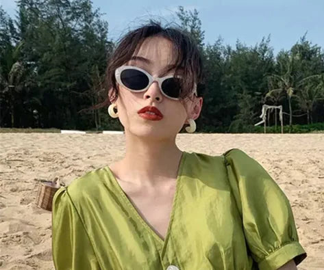 A woman wearing Retro Oval Sunglasses, red lipstick, and a shiny green dress poses on a sandy beach with trees in the background.