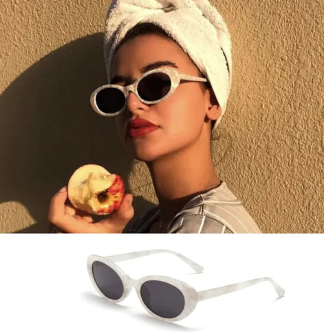 A woman wearing white retro oval sunglasses with a round frame, holding an apple. The sunglasses have a white frame and dark lenses.