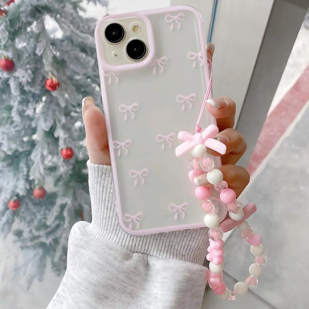 A pink iPhone 11 case with bow designs, accompanied by a matching pink and white beaded strap, held in a person's hand against a festive background.