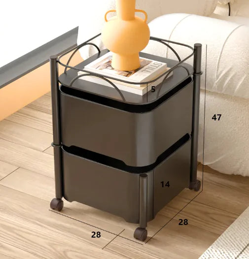 A black rotating snack storage rack with dimensions labeled on the image, placed on a wooden floor next to a bed with a yellow lamp on top.