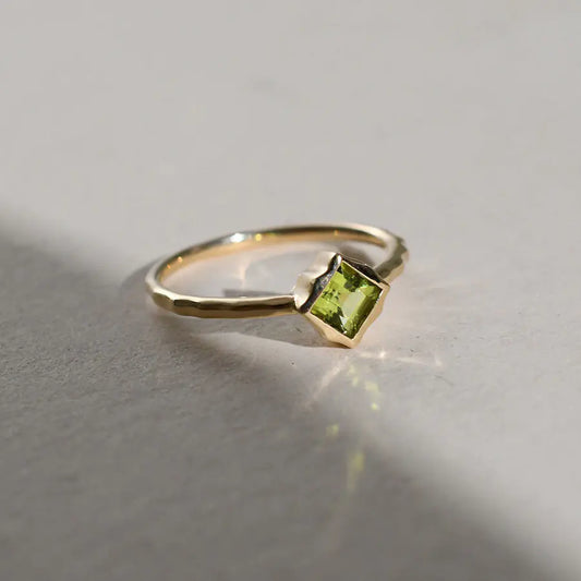 A silver ring with a square-cut green peridot stone, displayed against a neutral background with a source of light illuminating the stone.
