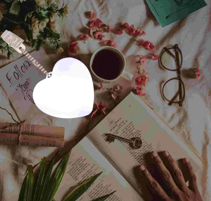 An LED automatic sensor purse light in a heart shape is illuminated and placed on an open book. The surrounding area includes a cup of coffee, glasses, and decorative flowers.