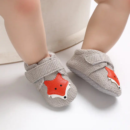 A pair of gray baby shoes with a cartoon fox design on the top, displayed on a baby's feet.