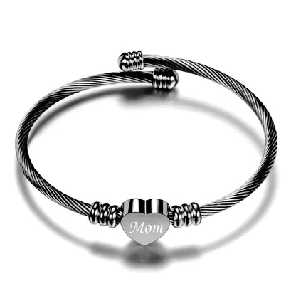 A stainless steel braided bracelet with a heart-shaped charm that has the word 'Mom' on it, displayed against a white background.