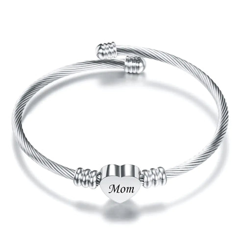 A stainless steel braided bracelet with a heart-shaped charm that has the word 'Mom' on it, displayed against a white background.