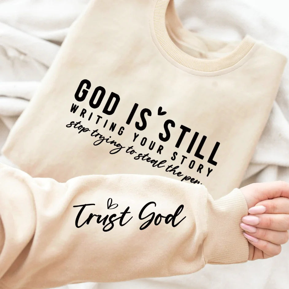 A gray crew neck sweatshirt with a text design 'GOD IS STILL WRITING YOUR STORY' printed on the front, paired with blue denim jeans.