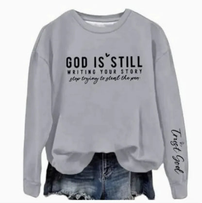 A gray crew neck sweatshirt with a text design 'GOD IS STILL WRITING YOUR STORY' printed on the front, paired with blue denim jeans.