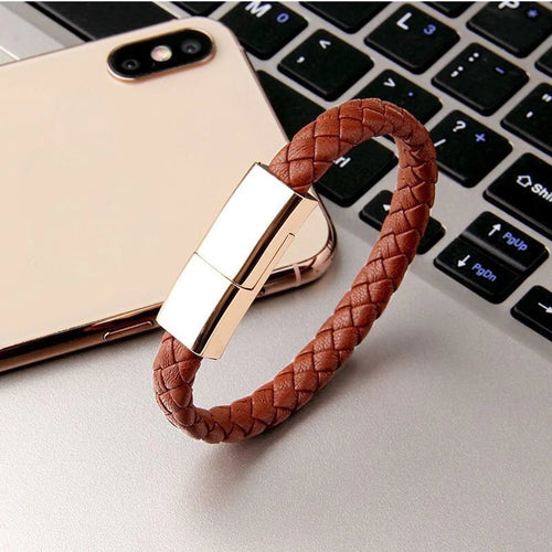 A bracelet charging cable designed to be worn on the wrist, displayed on a laptop next to a smartphone, showcasing its brown leather strap and gold-colored clasp.